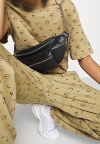 Černá kožená ledvinka se dvěma zipovými kapsami, nošená přes béžový outfit s kruhovým potiskem. Viditelné bílé tenisky s texturovanou podešví.