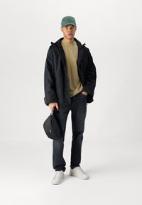 Svart vanntett jakke, olivengrønn t-skjorte, mørke jeans, hvite sneakers og en svart bag. Tilbehøret inkluderer en grønn caps.