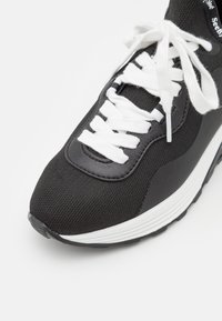 Chaussure de sport noire et blanche avec lacets blancs, bout en tissu mesh et semelle blanche épaisse sur un fond blanc uni.