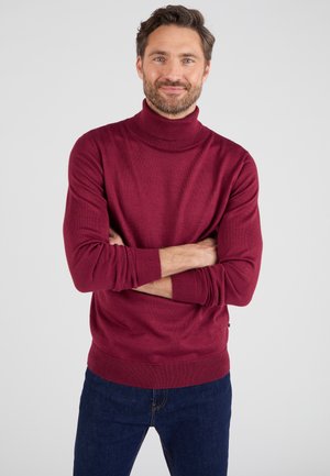 Maglione a collo alto color bordeaux in morbido tessuto a maglia, con colletto e polsini a coste, abbinato a jeans blu scuro, che mostrano una vestibilità rilassata.