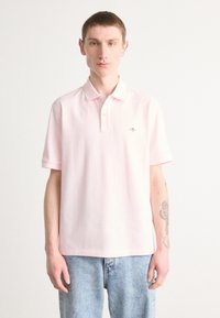 Polo en coton rose clair avec un col classique, des manches courtes et un petit logo brodé sur la poitrine. Porté avec un jean bleu.