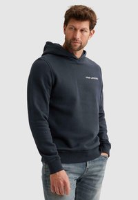 Hombre con una sudadera con capucha PME Legend de color azul oscuro y vaqueros azul claro, de pie con una mano en el bolsillo, mirando de lado contra un fondo liso.