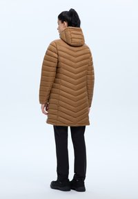 Bruine gewatteerde jas met een chevronpatroon, capuchon en lange mouwen. Draagt een zwarte broek en sneakers. Getextureerde stof met een gladde afwerking.