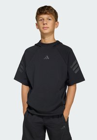 Ung person står med händerna i fickorna, klädd i en svart kortärmad luvtröja och svarta shorts med subtila Adidas-logotyper.