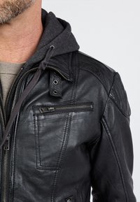 Zwarte leren jas met een ritssluiting aan de zak, een drukknopkraag en een grijze capuchonvoering. Gladde textuur met gedetailleerde stikaccenten.