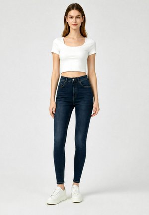 Donna in piedi con top corto bianco a maniche corte, jeans skinny blu scuro a vita alta e sneakers bianche con plateau su sfondo semplice.