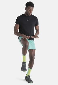 Camisa negra de manga corta, pantalones cortos verde menta y zapatos negros con acentos verdes; calcetines amarillo-verde brillante con un patrón texturizado.