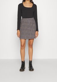 Person som bär en svart långärmad tröja, en marinblå och beige rutigt minikjol, och svarta snörda kängor, som står mot en slät vägg.