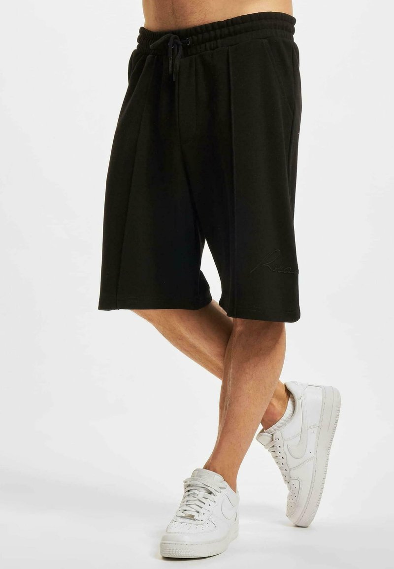 Black athletic shorts with an elastic waistband and drawstring, featuring two side pockets and embossed logo detail. Paired with white sneakers.