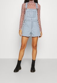 Overoles de denim azul claro con bolsillos frontales y tirantes ajustables, usados sobre una blusa de manga larga con estampado, combinados con botas negras con cordones.