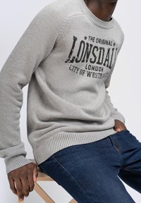 Gray knitted sweater with round neckline, featuring a distressed black print that reads “LONSDALE LONDON CITY OF WESTMINSTER.” Worn with blue jeans.