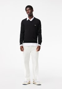 Maglione nero a scollo a v, indossato su una camicia bianca con colletto, abbinato a pantaloni bianchi e scarpe sportive casual. Logo verde ben visibile sul maglione.
