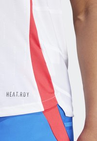 Biele športové dres s červeným bočným panelom, zdobené textúrovými vzormi a nápisom "HEAT.RDY" vytlačeným na leme. Dopĺňané modrými šortkami.