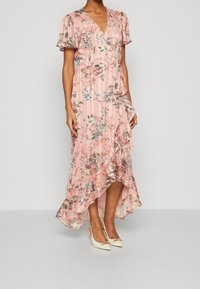 Blomstret maxikjole i blød pink stof med flagrende ærmer, en V-hals og asymmetrisk kant. Et par hvide hæle med guld accenter.