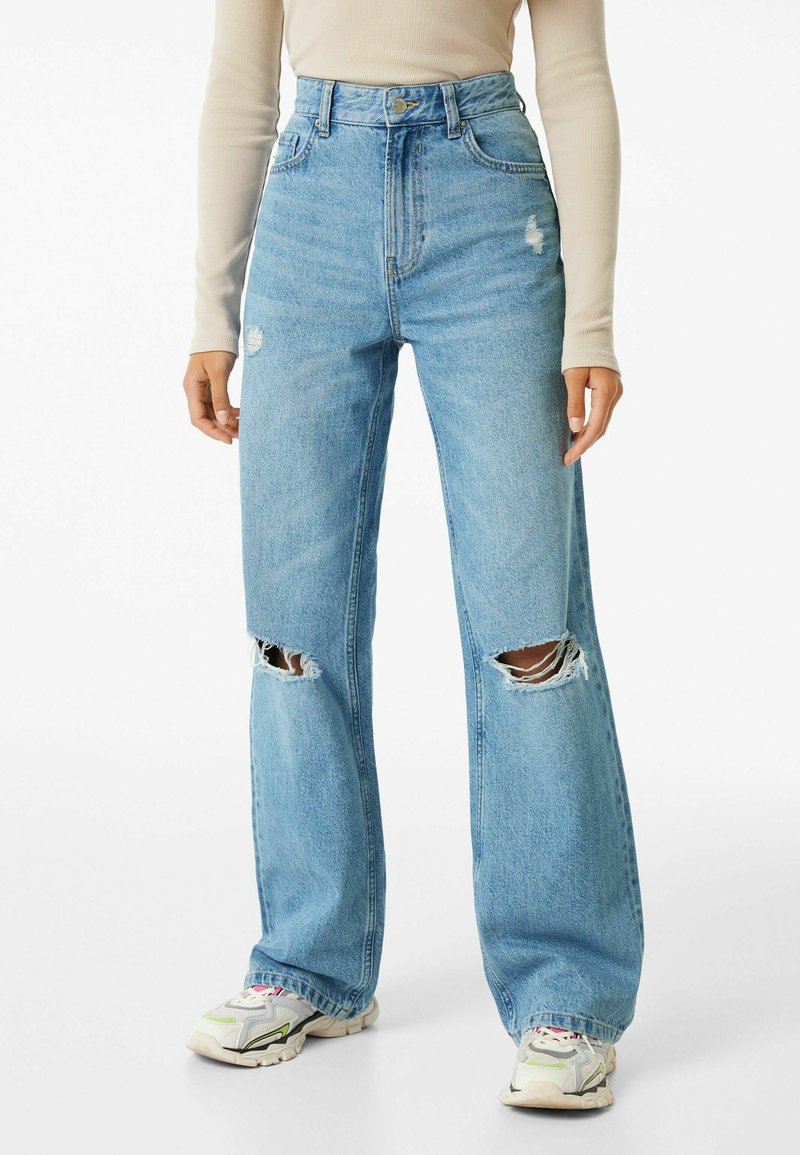 Person med högmidjade ljusblå slitna jeans med hål vid knäna, beige långärmad topp och vita sneakers.