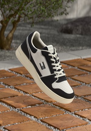 Schwarzer und weißer Sneaker mit weißen Schnürsenkeln, schwebend über einem Holzblock und einer Kiesfläche im Freien mit einem Baum im Hintergrund.