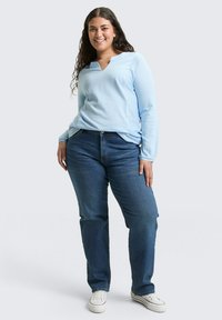 Jasnoniebieska bluzka z długim rękawem w pionowe paski, z dekoltem w kształcie V i luźnym krojem, zestawiona z ciemnoniebieskimi dżinsami prostymi i białymi trampkami.