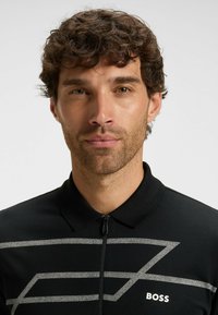 Czarna koszulka polo z kołnierzykiem na zamek, z odważnym srebrnym geometrycznym wzorem oraz nazwą marki "BOSS" w białym kolorze na dole.