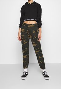 Svart cropped hoodie med en tjock banderoll med texten "URBAN CLASSICS." Camo-byxor i gröna och bruna nyanser, med textur och cargofickor.