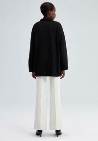 Vrouw met kort haar in een zwarte oversized trui, witte wijde broek en zwarte hakken, die met de rug naar de camera staat op een lichtgrijze achtergrond.