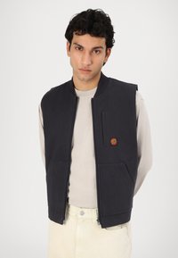 Dark blue sleeveless vest featuring a front zipper, two front pockets, and a small red logo patch. Worn over a light cream shirt.