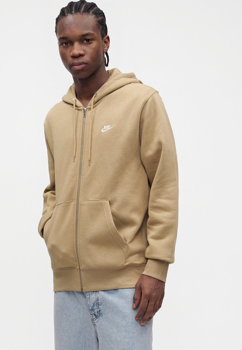 Young man with braided hair wearing a tan Nike zip-up hoodie and light blue jeans against a plain white background.