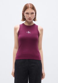 Kurz geschnittenes, ärmelloses Oberteil aus burgunderrotem Rippstoff mit rundem Ausschnitt und kleinem weißem Logo in der Mitte der Brust. Kombiniert mit dunklen Hosen.