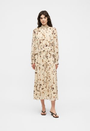 Woman wearing a long, beige floral dress with long sleeves, high neck, ruffle detail at waist, and black heeled sandals, standing on a white background.