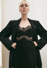 Vestido de satén negro con detalles de encaje en el cuerpo, superpuesto bajo una blazer negra entallada con mangas anchas. Textura suave, diseño elegante.