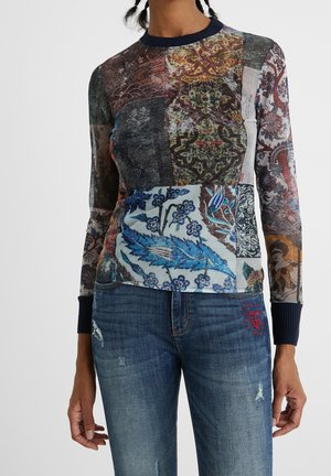 Haut ajusté à manches longues avec motifs floraux et abstraits multicolores de style patchwork, associé à un jean bleu avec broderie cœur rouge.