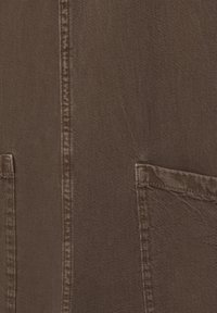 Primo piano di un tessuto marrone con cuciture verticali e due tasche visibili, che mette in risalto la texture e i dettagli delle cuciture.