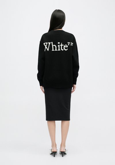 Maglione nero a maglia con il testo bianco "White™" sulla schiena, caratterizzato da maniche lunghe e una vestibilità rilassata. Abbinato a una gonna nera a tubino.