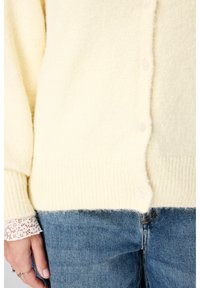 Lichtgele gebreide cardigan met een zachte textuur, voorzien van ronde plastic knopen, geribde manchetten en zoom, gedragen over een wit shirt met kanten rand.