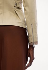 Gros plan d'une personne portant une veste beige avec une fermeture éclair sur la manche et une jupe marron sur fond blanc.