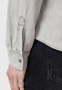 Helles graues Denim-Hemd mit einem knöpfbaren Manschette und einem dunklen Knopf, kombiniert mit dunklen Denim-Hosen. Strukturiertes Gewebe und taillierte Passform.