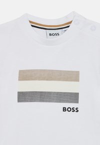 Λευκό βαμβακερό μπλουζάκι με οριζόντιο ρίγες σε καφέ, κρεμ και γκρι. Το λογότ υπο "BOSS" εκτυπωμένο με μαύρο κάτω από το σχέδιο.