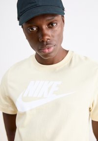 Camiseta de algodón amarillo claro con un prominente logo blanco de Nike. El modelo lleva una gorra oscura, destacando el contraste de color de la camiseta.