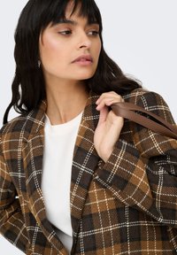 Sacou în carouri maro și negru dintr-un material texturat, cu rever ascuțit și căptușeală dublă ton. Top alb dedesubt, ținând o curea maro de geantă.