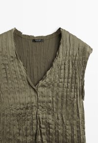 Blusa verde oliva senza maniche con scollo a V profondo e tessuto arricciato. Presenta un orlo grezzo e chiusura frontale con bottoni.