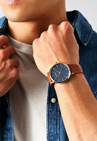 Brauner Lederarmbanduhr mit blauem Zifferblatt, goldenen Akzenten und rundem Gehäuse. Am Handgelenk getragen, zeigt sie die Hände und das Hemd unter einer Jeansjacke.