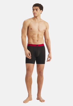 Uomo muscoloso in piedi a piedi nudi, indossa boxer neri con una fascia in vita rossa e il logo Adidas, guarda di lato su sfondo bianco.