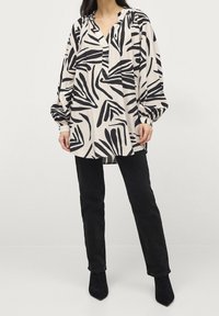 Langærmet bluse i creme med sort abstrakt mønster, med en placket-krave og gatherede ærmer, parret med sorte jeans.