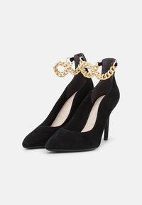 Svarta mocka högklackade pumps med spetsig tå och en ankelrem prydd med en guldkedjedetalj. Slät textur, elegant design.