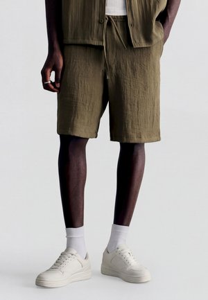 Muž nosiaci olivovo zelené textúrované kraťasy, biele ponožky a biele tenisky, stojaci s jednou rukou popri tele a druhou v vrecku.