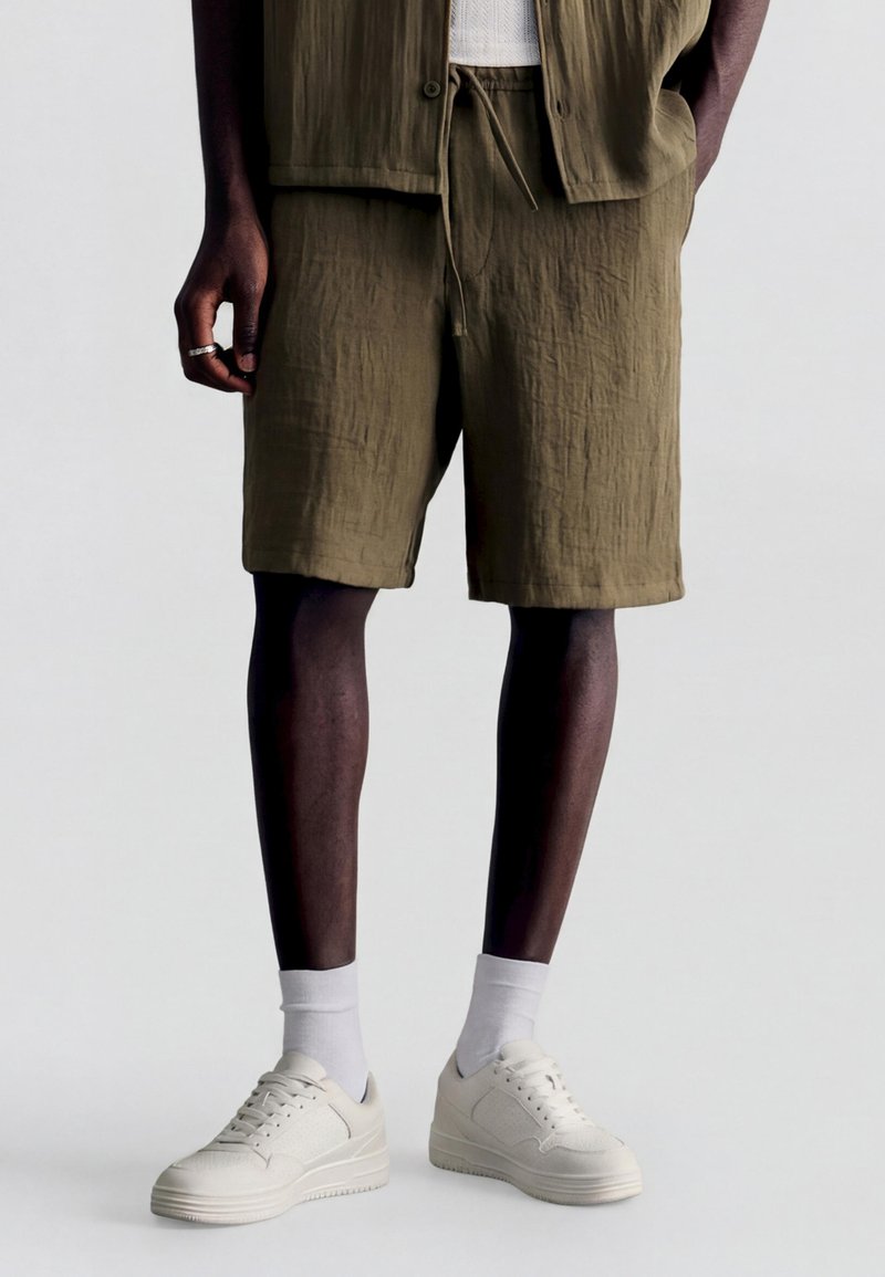 Homme portant un short texturé vert olive, des chaussettes blanches et des baskets blanches, debout avec une main le long du corps et l’autre dans la poche.