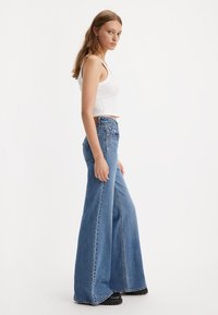 Noor noor valges ilma varrukateta kroptopist ja kõrge vöökohaga laia jalaga sinistes teksades, seistes valge tausta ees.