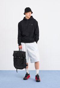 Tânăr îmbrăcat cu un hanorac negru, pălărie tip bucket, pantaloni scurți albi și adidași roșii, ținând un rucsac negru, pe un fundal simplu.