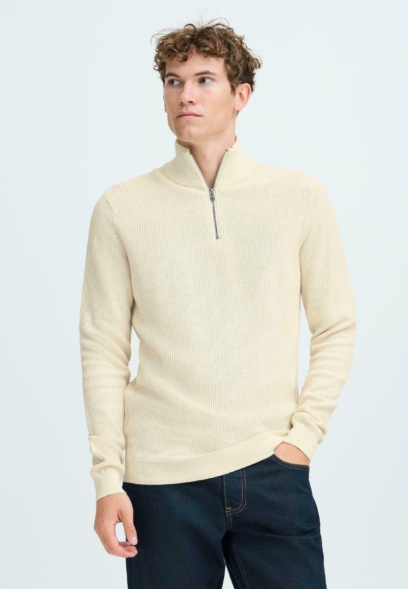Beige ritsvest met een textuurgebreid ontwerp, lange mouwen en ribgebreide manchetten. Gecombineerd met donkere jeans voor contrast.