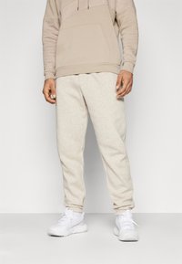 Beige sweatshirt met een gestructureerd bovenpaneel, zak aan de voorkant en een trekkoord. Bijpassende beige joggingbroek met een zachte textuur en elastische boorden. Witte schoenen.