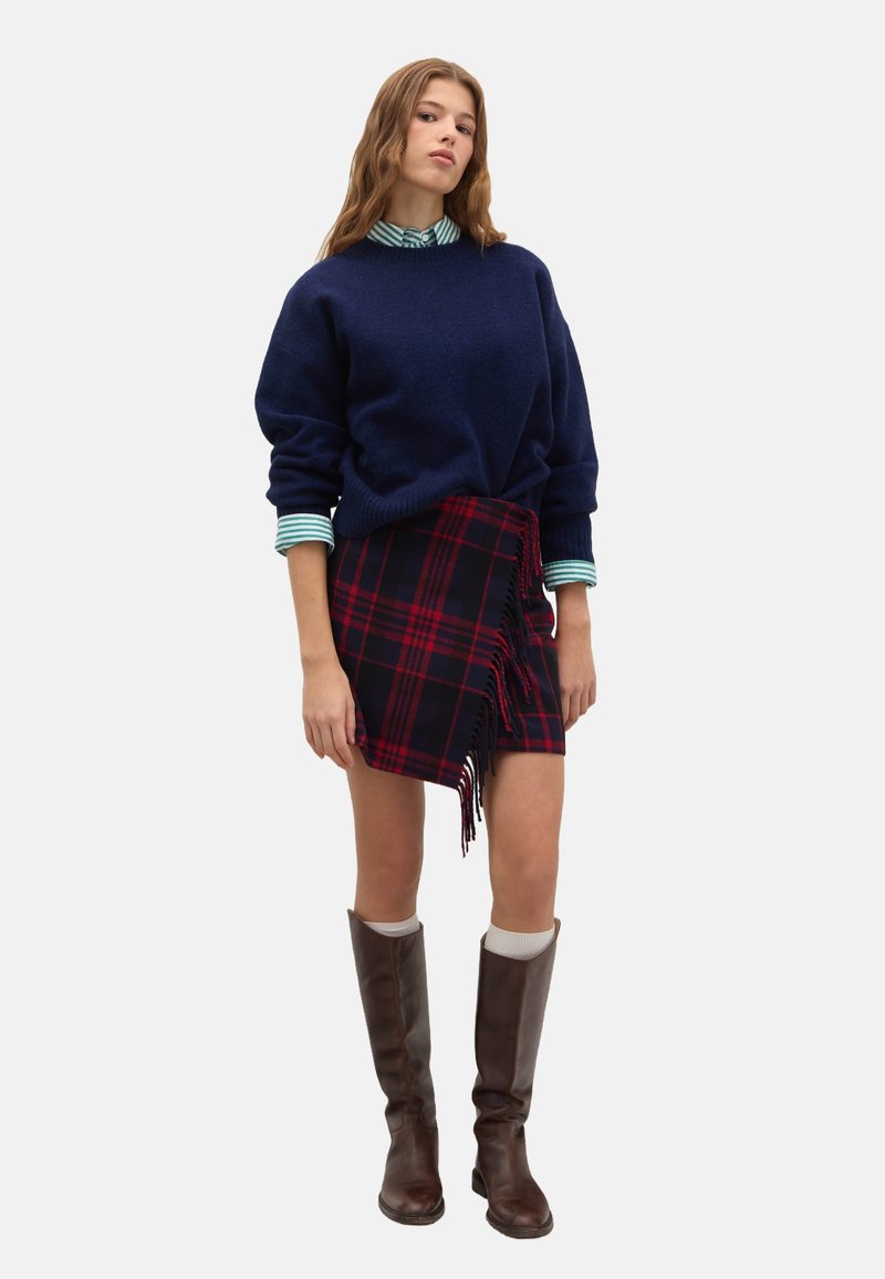 Un suéter de gran tamaño en azul marino combinado con una falda de cuadros roja y negra con bordes deshilachados. Botas marrones hasta la rodilla completan el atuendo.